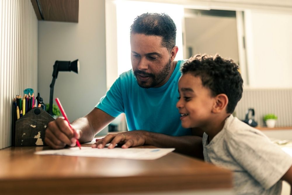 An adult and a child sit at a desk together. The adult writes on a paper with a red pen while the child smiles and watches. Various colored pencils are visible on the desk beside them.