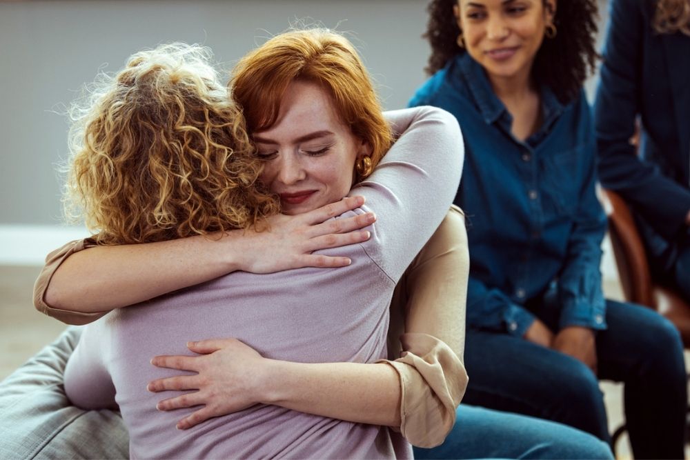 Two women embrace warmly in the foreground, while another woman sits nearby, smiling and observing their interaction.