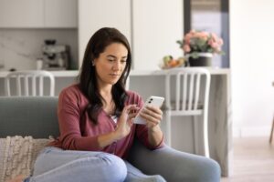 A woman sits on a couch in a modern kitchen, looking at and using her smartphone. She wears a pink cardigan and blue jeans, with a pillow and flowers visible in the background.