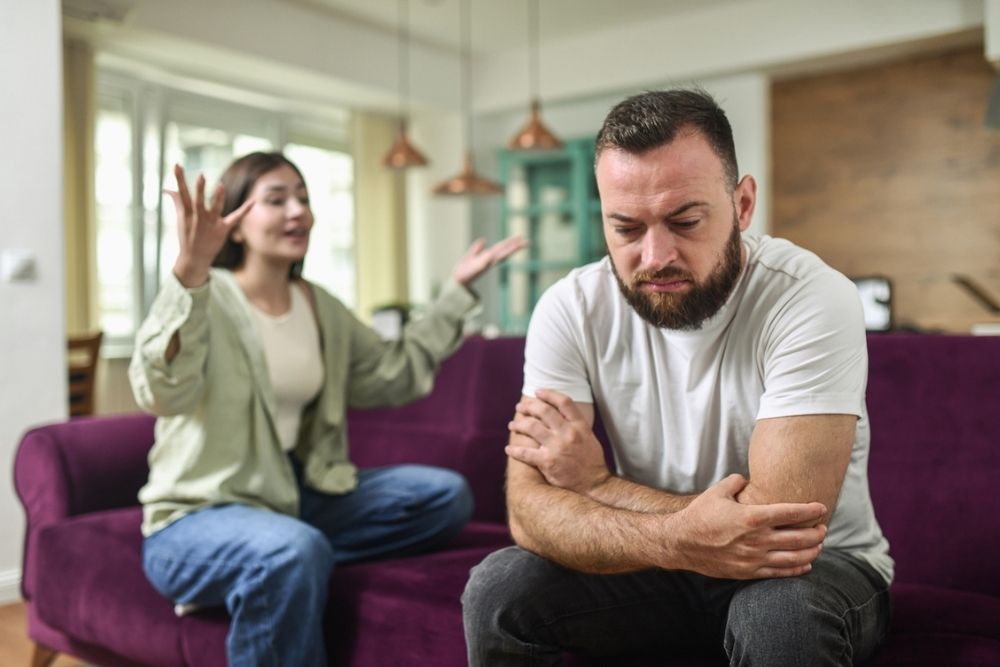 A woman gestures emotionally while talking to a man sitting on a purple couch. The man looks upset with arms crossed, avoiding eye contact. They appear to be having a serious conversation in a living room.