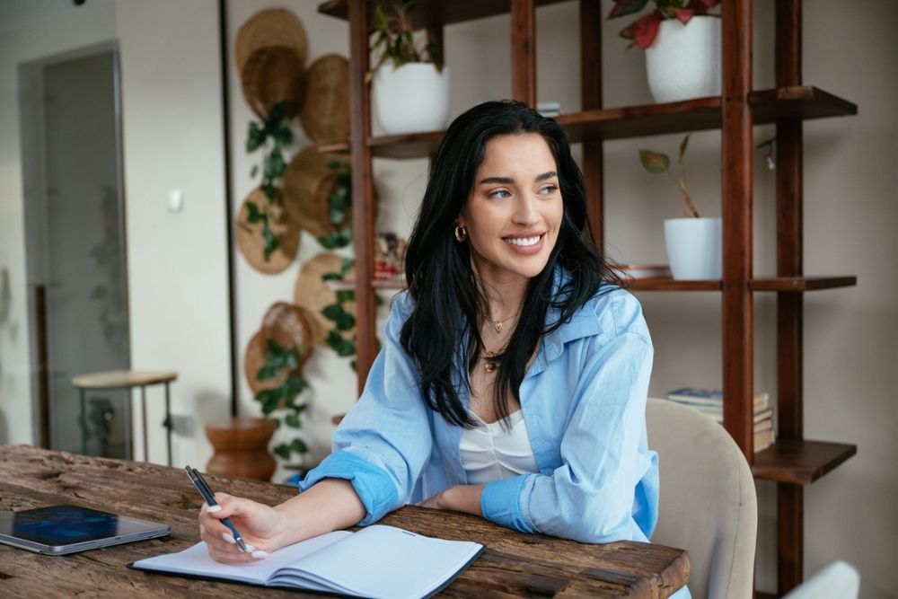 A woman with long dark hair sits at a wooden table, smiling and holding a pen over an open notebook. A tablet is nearby, and there are shelves with plants and hats in the background.