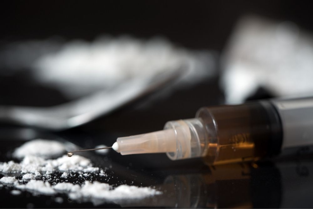 A syringe with a droplet at the needle tip lies next to piles of white powder and a metal spoon, all on a dark surface.