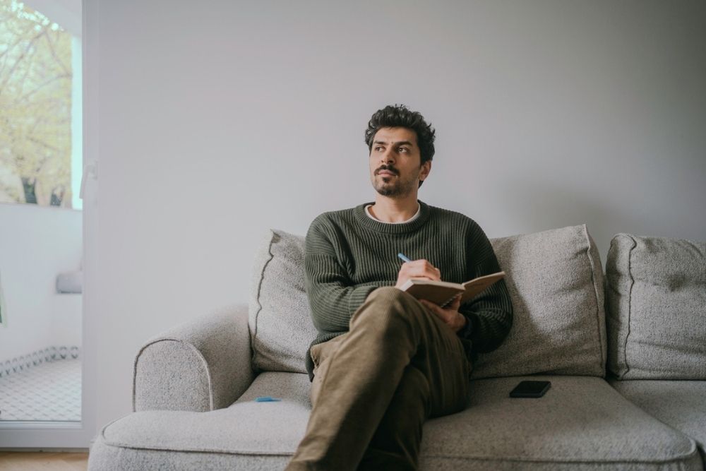 A man sits on a gray couch, holding a notebook and pen, looking thoughtfully upward. A smartphone lies beside him on the couch, and light comes in from a nearby window.