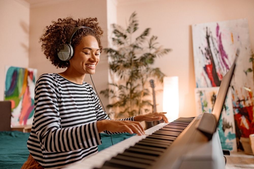 A person wearing headphones and a striped shirt smiles while playing a keyboard piano in a room with houseplants and colorful abstract paintings in the background.