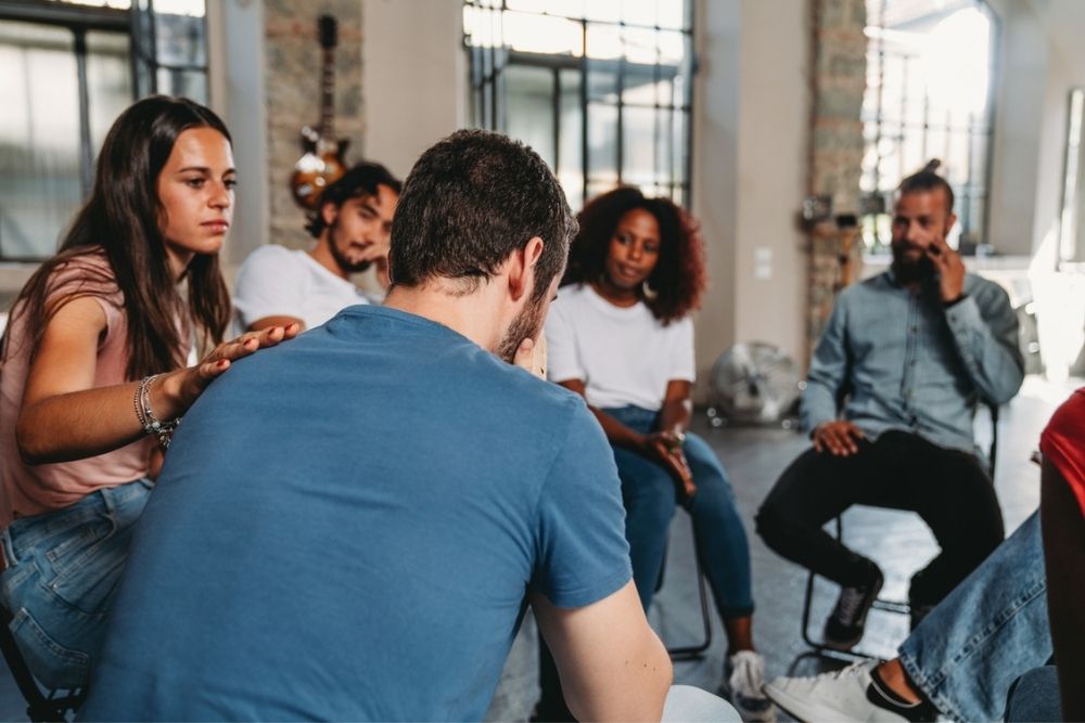 A woman comforts a man by placing a hand on his back during a group therapy session. Other people sit in a circle, listening attentively in a bright room.