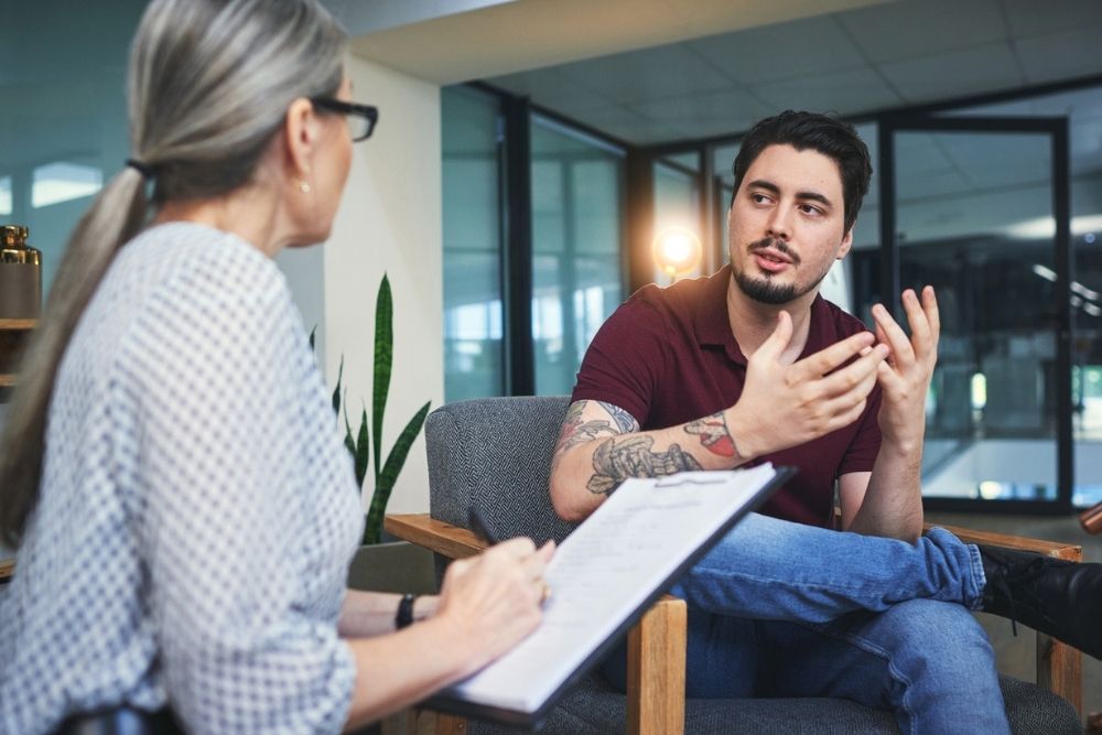 A man with tattoos is talking expressively to a woman holding a clipboard during a counseling or therapy session in a modern office.