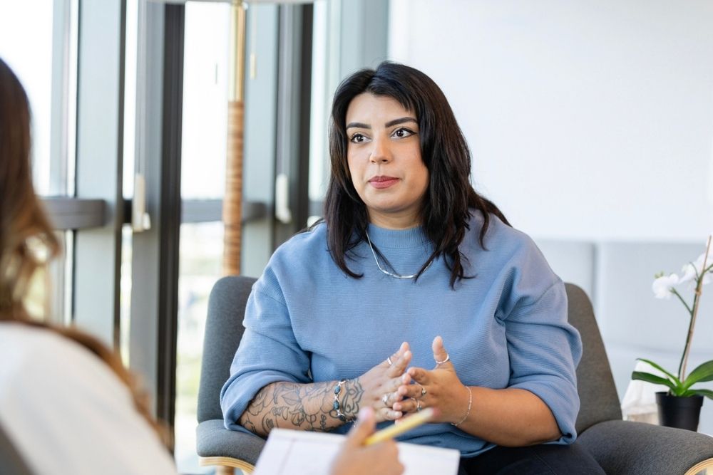 A woman with dark hair and tattoos, wearing a blue top, sits in a chair talking to another person whose back is visible, in a bright room with large windows.