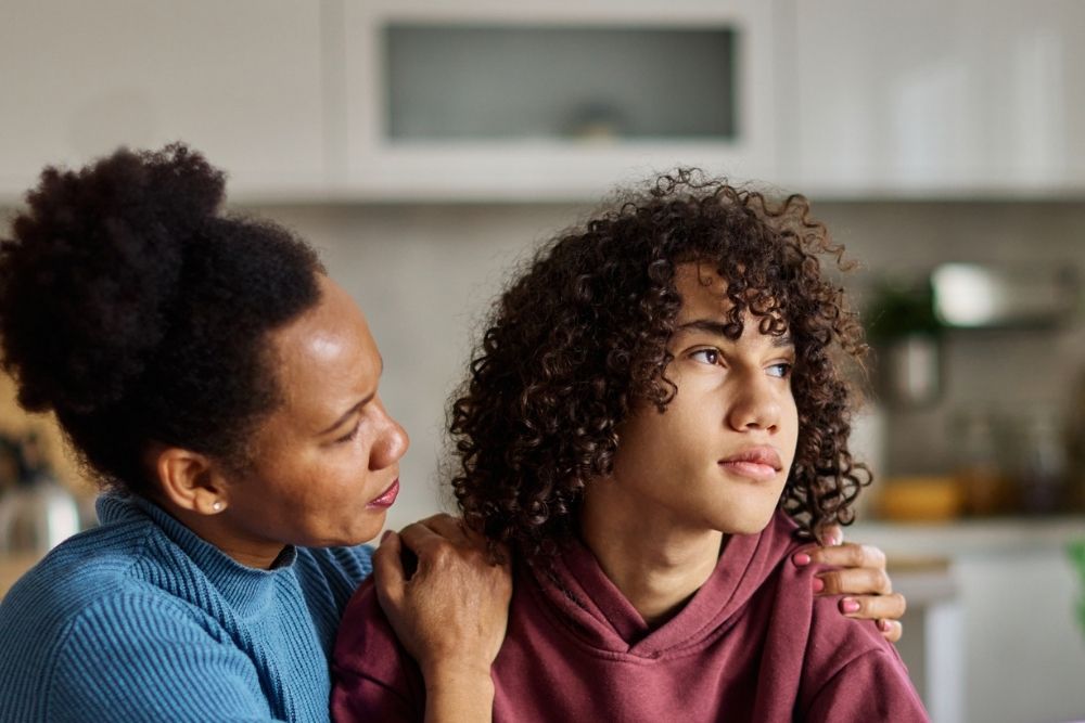 A woman gently holds a teenagers shoulders, offering comfort. The teen looks away with a distant expression, while the woman appears concerned. They are indoors in a kitchen setting.