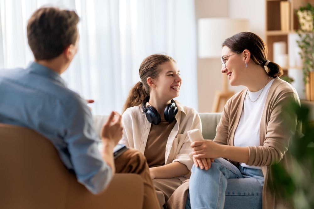 A teenage girl with headphones around her neck smiles at an adult woman beside her, while another adult faces them, holding a notebook in a bright, comfortable room.