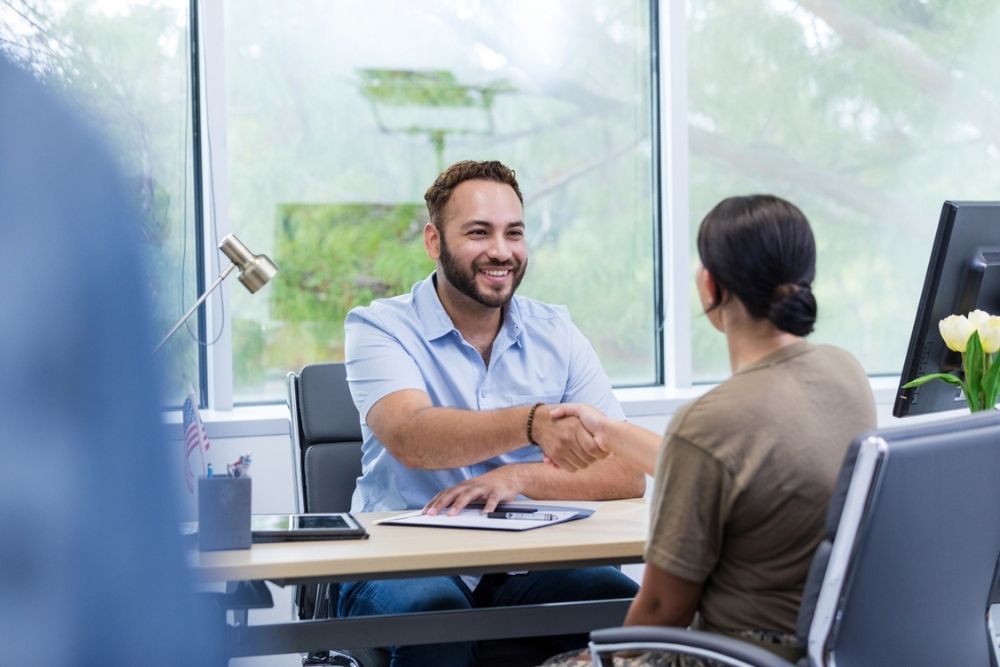 A man and a woman sit across from each other at an office desk, smiling and shaking hands. The office has large windows with greenery outside and a computer, notebook, and flowers on the desk.